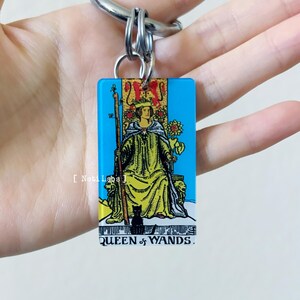 May include: A blue and yellow keychain featuring the Queen of Wands tarot card. The card is surrounded by a blue border and the text "QUEEN of WANDS" is printed at the bottom.