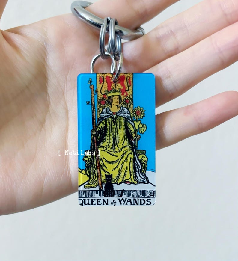 May include: A blue and yellow keychain featuring the Queen of Wands tarot card. The card is surrounded by a blue border and the text "QUEEN of WANDS" is printed at the bottom.