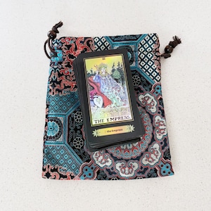 May include: A deck of tarot cards with "THE EMPRESS" printed on the top card rests on a patterned drawstring bag. The bag features a teal, red, and black design. The card has a gold border and depicts a woman.
