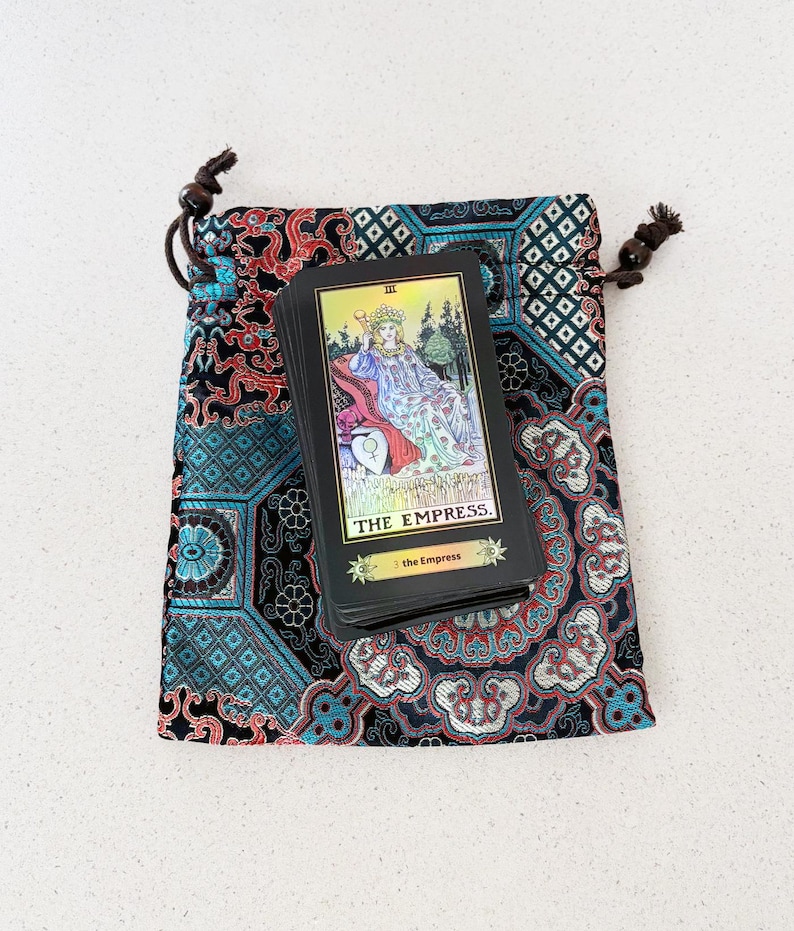 May include: A deck of tarot cards with "THE EMPRESS" printed on the top card rests on a patterned drawstring bag. The bag features a teal, red, and black design. The card has a gold border and depicts a woman.