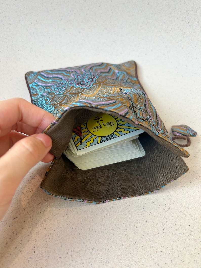 Puede incluir: Una baraja de tarot con una bolsa de patr&oacute;n dorado y azul. La carta superior es la carta del Sol, que es la 19&ordf; carta del Arcano Mayor.