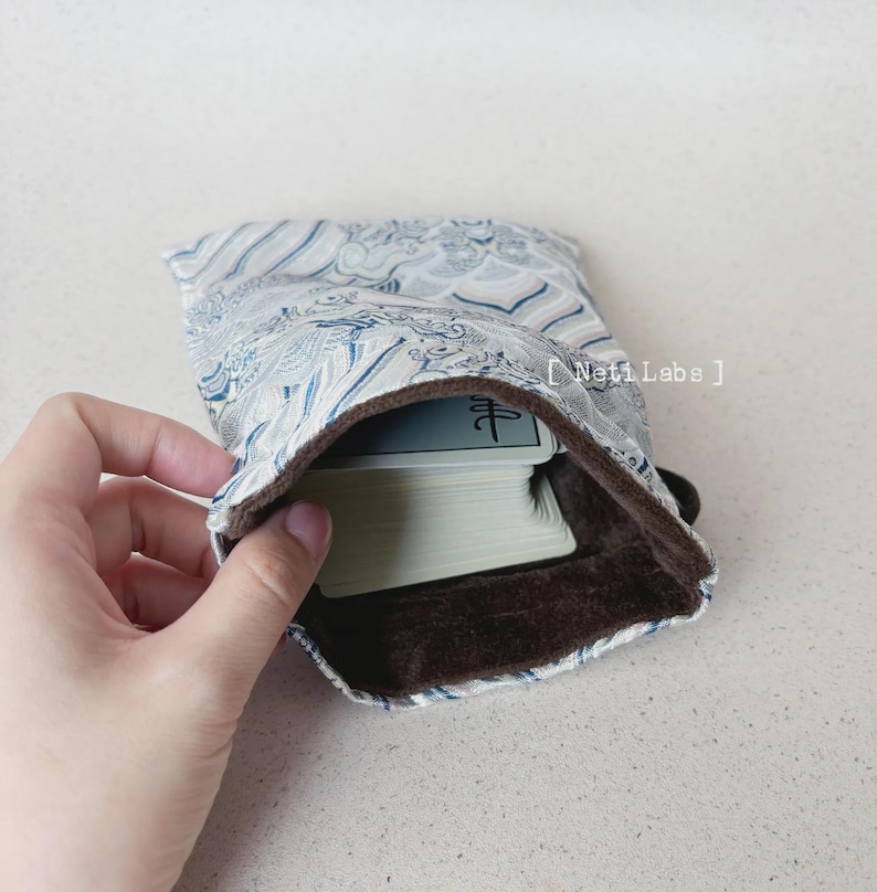 Puede incluir: Una bolsa de tela con estampado blanco y azul con forro de terciopelo marr&oacute;n. La bolsa est&aacute; abierta y dentro hay una baraja de cartas. La carta superior tiene un solo car&aacute;cter chino.