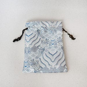 Puede incluir: Una peque&ntilde;a bolsa de cord&oacute;n blanco con un dise&ntilde;o estampado azul y blanco con nubes y dragones. La bolsa tiene cordones de cord&oacute;n negros con cuentas de madera.