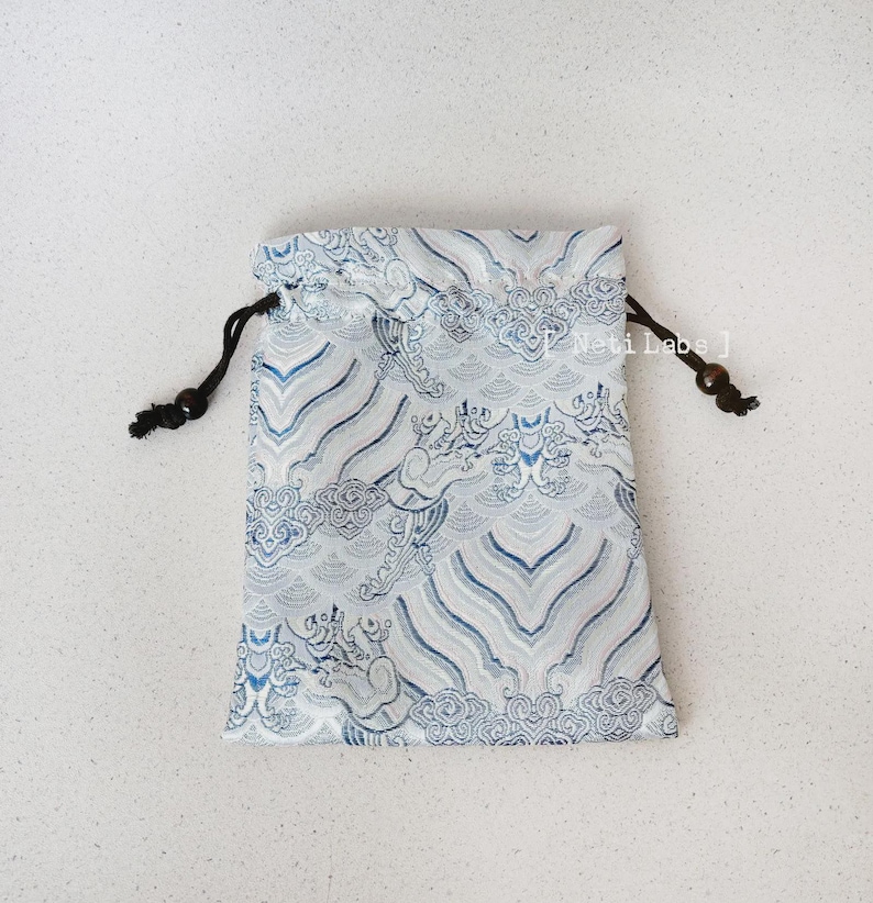 Puede incluir: Una peque&ntilde;a bolsa de cord&oacute;n blanco con un dise&ntilde;o estampado azul y blanco con nubes y dragones. La bolsa tiene cordones de cord&oacute;n negros con cuentas de madera.