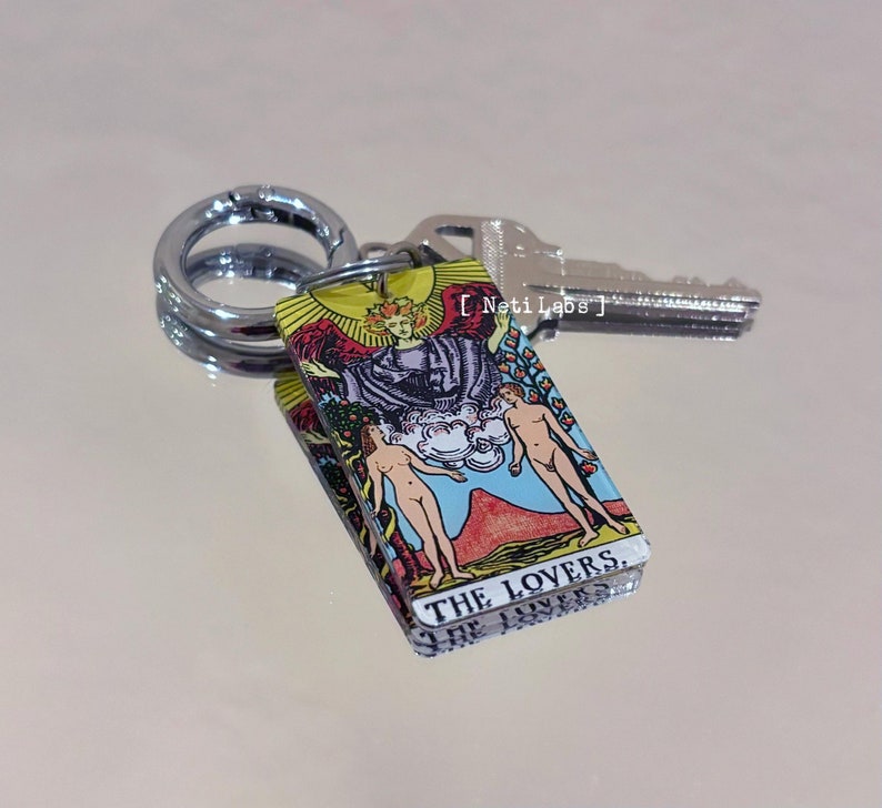 May include: A keychain featuring a yellow card with a colorful illustration of The Lovers tarot card. The card has the text "THE LOVERS" printed on it. The keychain is attached to a silver key ring.