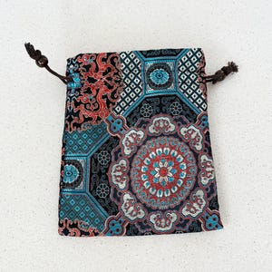 May include: A small, patterned drawstring bag. The bag features a detailed design with teal, red, and white floral and geometric patterns on a black background. The drawstring is brown.
