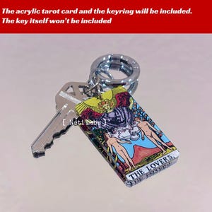 May include: A silver keychain with a clear acrylic charm featuring the Lovers tarot card. The card has a colorful illustration of a couple standing in a garden with a winged figure above them. The text "THE LOVERS" is printed on the bottom of the card.