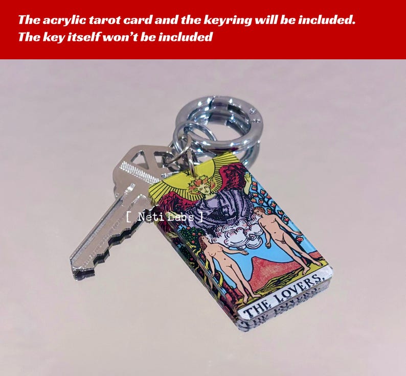 May include: A silver keychain with a clear acrylic charm featuring the Lovers tarot card. The card has a colorful illustration of a couple standing in a garden with a winged figure above them. The text "THE LOVERS" is printed on the bottom of the card.