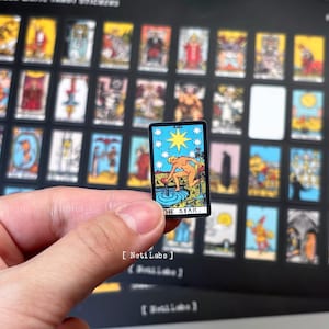 May include: A close-up of a tarot card with a woman pouring water from a pitcher into a pool. The card is titled "The Star" and has a blue, white, and yellow color scheme.
