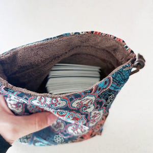 May include: A patterned drawstring bag with a stack of cards inside. The bag features a blue, red, and white floral design on a dark background. The interior is lined with brown, soft material. A brown leather cord is used to close the bag.