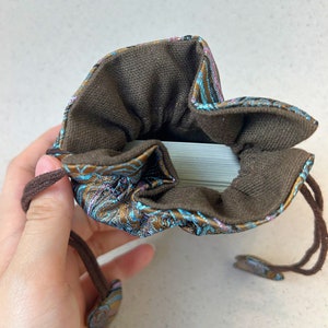Puede incluir: Una bolsa de cord&oacute;n marr&oacute;n con un patr&oacute;n floral azul y dorado. La bolsa est&aacute; abierta y se puede ver una baraja de cartas en su interior.