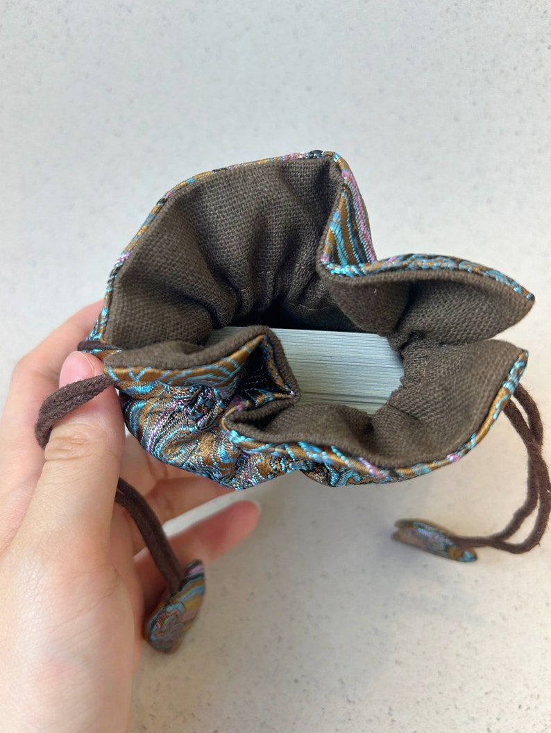 Puede incluir: Una bolsa de cord&oacute;n marr&oacute;n con un patr&oacute;n floral azul y dorado. La bolsa est&aacute; abierta y se puede ver una baraja de cartas en su interior.