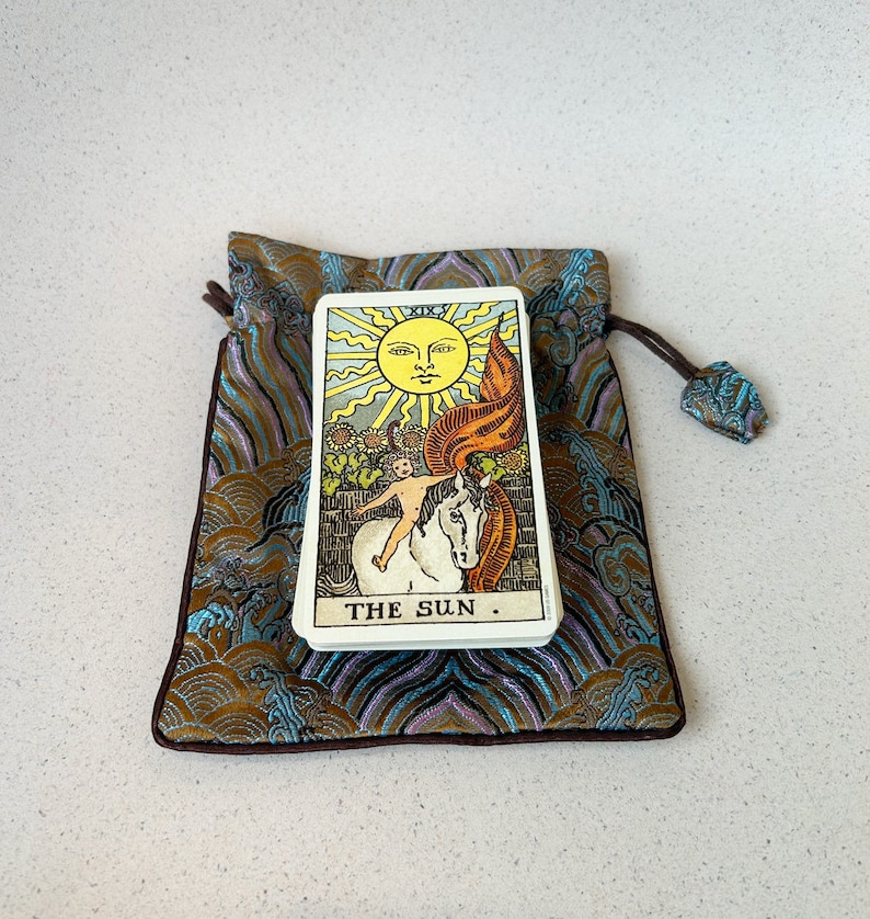 Puede incluir: Una carta del tarot con un sol amarillo y una persona montando un caballo blanco. La carta se llama "El Sol" y est&aacute; en una bolsa azul y dorada.