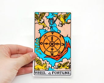 May include: Tarot card depicting the Wheel of Fortune. The card features a wheel with symbols and a sphinx riding a red creature. The card is set against a blue background with clouds and two winged creatures.