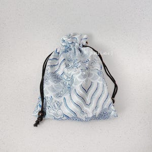 Puede incluir: Una bolsa de cord&oacute;n blanco con un dise&ntilde;o estampado azul y plateado. La bolsa tiene un cord&oacute;n negro y una peque&ntilde;a cuenta de madera en el extremo de cada cuerda.