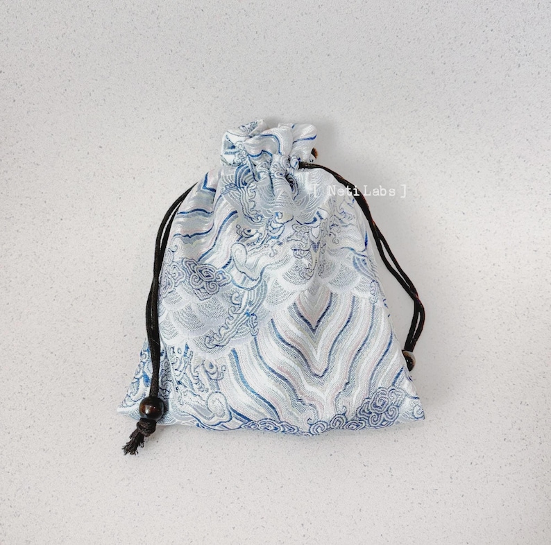 Puede incluir: Una bolsa de cord&oacute;n blanco con un dise&ntilde;o estampado azul y plateado. La bolsa tiene un cord&oacute;n negro y una peque&ntilde;a cuenta de madera en el extremo de cada cuerda.