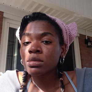 May include: A person wearing a light purple crocheted cat ear headband, silver hoop earrings, and a silver necklace with a pendant. The person is wearing a gray top with a blue undershirt.