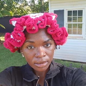 May include: A handmade crochet head covering in a vibrant fuchsia and white colour scheme. The head covering features a unique design with multiple flower-like embellishments. The person in the image is wearing a black jacket.