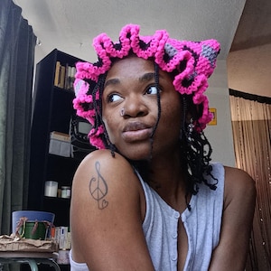 May include: A handmade, pink and grey crocheted head covering with a ruffled edge. The head covering is worn by a person wearing a grey vest top. A tattoo of a musical note and peace sign is visible on the person's shoulder.