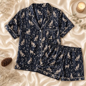 Könnte beinhalten: Ein dunkelblaues Pyjama-Set bestehend aus einem kurzärmeligen Hemd mit Knopfleiste und passender Shorts. Der Stoff zeigt ein durchgehendes Muster aus weißen Füchsen, Sternen und kleinen Blumen, verziert mit einer feinen weißen Paspelierung.
