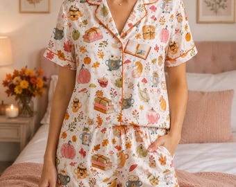 Könnte beinhalten: Ein weißer kurzärmeliger Pyjama mit einem orangefarbenen, geknöpften Oberteil und passender Shorts, verziert mit einem wiederkehrenden Muster aus herbstlichen Motiven wie Kürbissen, Bücherstapeln, Tassen mit Heißgetränken und Laub.