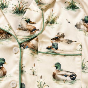 May include: A cream-colored button-down shirt featuring a repeating print of mallard ducks swimming in water with green reeds. The garment has a notched collar and a chest pocket, both accented with thin green piping along the edges.