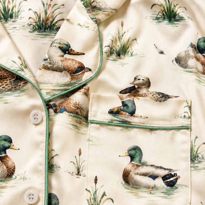 May include: A cream-colored button-down shirt featuring a repeating print of mallard ducks swimming in water with green reeds. The garment has a notched collar and a chest pocket, both accented with thin green piping along the edges.