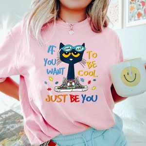 Puede incluir: Camiseta rosa con un gato negro que lleva gafas de sol que dicen "Cool Cat". El gato lleva un par de zapatos con un patrón de piezas de rompecabezas. El texto en la camiseta dice "Si quieres ser cool, solo sé tú mismo".