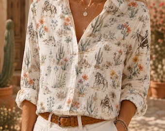 Puede incluir: Una camisa de manga larga con botones en tela color hueso con un estampado de temática desértica que incluye ilustraciones de cactus, flores silvestres coloridas y figuras a caballo. La camisa está metida en un pantalón blanco con cinturón.