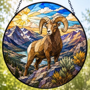 May include: A circular stained glass suncatcher featuring a bighorn sheep standing on a rocky ledge. The background depicts a mountain landscape with a lake, pine trees, and a bright yellow sun in a blue sky. The piece is framed in black metal.