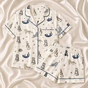 May include: A two-piece pajama set featuring a short-sleeved button-down shirt and matching shorts. The fabric is cream-colored with a repeating pattern of various cats in shades of gray, brown, and black, accented by dark blue piping trim.