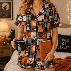 May include: A short-sleeve button-down pajama shirt and matching shorts set featuring a patchwork pattern of orange, black, and white Halloween motifs including ghosts, pumpkins, bats, and floral designs. A black mug with the text HEY BOO is held.