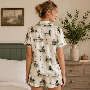 May include: A short-sleeved button-down shirt and matching shorts set in a cream-colored fabric featuring an all-over print of mallard ducks swimming in water with green reeds. The garment has dark green piping along the collar and edges.