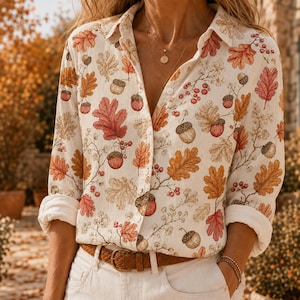 May include: A long-sleeved button-down shirt in an off-white color featuring an all-over print of autumn-themed oak leaves, acorns, and small red berries. The shirt has a classic collar, a front button placket, and cuffs that are rolled up.