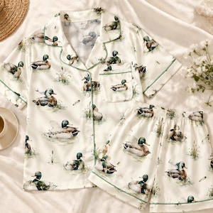 May include: A cream-colored pajama set featuring a short-sleeve button-down shirt with a notched collar and a chest pocket, paired with matching elastic-waist shorts. The fabric is decorated with a repeating pattern of mallard ducks in water.