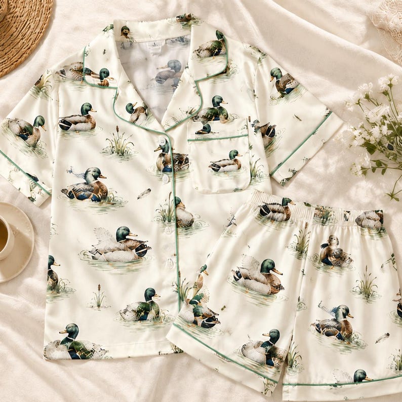 May include: A cream-colored pajama set featuring a short-sleeve button-down shirt with a notched collar and a chest pocket, paired with matching elastic-waist shorts. The fabric is decorated with a repeating pattern of mallard ducks in water.