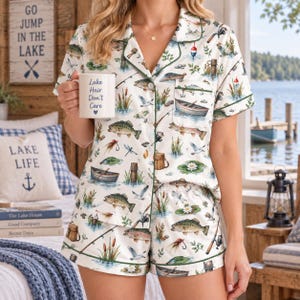 Puede incluir: Un conjunto de pijama blanco de manga corta con botones y pantalón corto a juego, con un estampado colorido de peces, cañas de pescar, barcas y nenúfares. La camisa tiene cuello con solapa y ribete verde, y una taza blanca con el texto Lake Hair Don't Care.