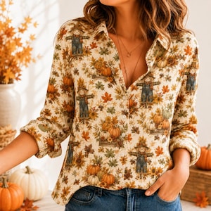 May include: A long-sleeved button-down shirt featuring an all-over print of scarecrows, pumpkins, and autumn leaves in shades of orange, brown, and green on a cream background. The shirt has a classic collar and a front button closure.
