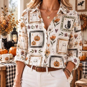 May include: A long-sleeve button-down shirt featuring a cream background with a repeating pattern of framed illustrations including ghosts, jack-o'-lanterns, black cats, spiderwebs, and orange flowers, paired with a brown leather belt and white pants.