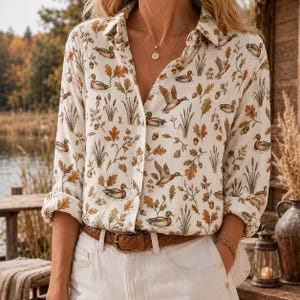 Peut inclure: Une chemise à manches longues boutonnée avec un imprimé intégral de canards colverts, de feuilles de chêne et de glands sur un fond crème. La chemise présente un col classique, une patte de boutonnage et est rentrée dans un pantalon clair avec une ceinture marron.