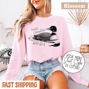 Può includere: Felpa rosa chiaro con grafica nera di un'uccello tuffatore nell'acqua, con la scritta "Stupid Canadian Wolf Bird". La parola "Blossom" è in alto a destra. Una grafica circolare dice "Pick More Shirt Styles In Listing!"