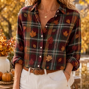 May include: A long-sleeved button-down shirt featuring a plaid pattern in shades of dark red, green, and yellow, accented with a scattered print of autumn leaves. The shirt is worn tucked into white pants with a brown leather belt.