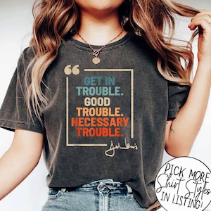Op de afbeelding: Een donkergrijs T-shirt met een vintage-stijl grafische afbeelding met de tekst "Get in Trouble. Good Trouble. Necessary Trouble." met een handtekening eronder. De afbeelding bevindt zich in een wit kader met een regenboogkleurenschema.