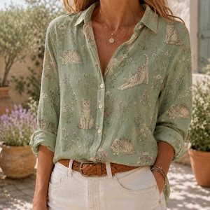 May include: A light green long-sleeve button-down shirt featuring a repeating pattern of cats and wildflowers. The shirt has a classic collar, white buttons down the front, and is tucked into white pants with a brown braided leather belt.