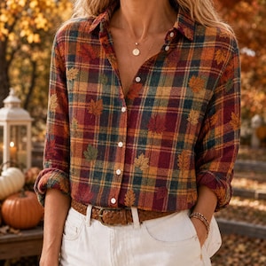 May include: A long-sleeve button-down shirt featuring a plaid pattern in shades of red, orange, yellow, and green, accented with small embroidered maple leaf motifs. The shirt is tucked into white trousers with a brown leather belt.