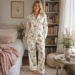 May include: A two-piece pyjama set consisting of a long-sleeved button-down shirt and matching wide-leg trousers. The fabric is off-white and features a repeating pattern of stacked books, wildflowers, and green foliage with thin pink piping.