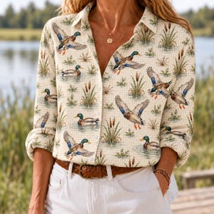May include: A long-sleeve button-down shirt featuring a repeating pattern of mallard ducks in flight and swimming among cattails and reeds. The shirt has a light beige background with a subtle grid texture and a classic collar with white buttons.