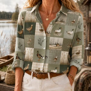 May include: A long-sleeve button-down shirt in a muted sage green and beige patchwork pattern featuring various illustrations of ducks in natural settings, including reeds and trees, with a classic collar and a brown leather belt at the waist.