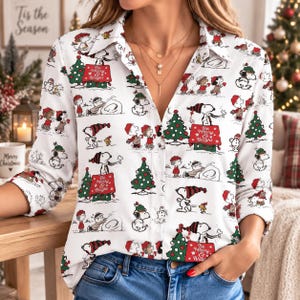 May include: A white long-sleeve button-down shirt featuring a repeating pattern of Snoopy and Charlie Brown characters in winter attire, Christmas trees, and red signs with the text Be Merry & Bright, paired with blue denim jeans.