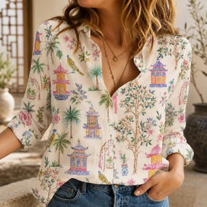 May include: A long-sleeved button-down shirt in an off-white color featuring a colorful print of pagodas, palm trees, flowering trees, and birds. The shirt has a classic collar, a front button placket, and cuffs that are rolled up to the elbows.